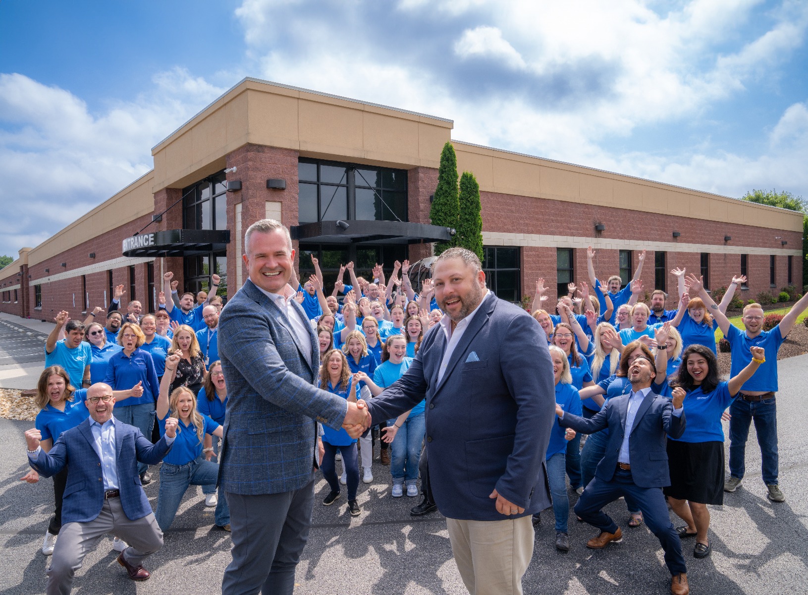 Christopher Gaines and Erik Miller shaking hands in a parking lot in front of the Inorganic Ventures building with a large group of people celebrating behind them.